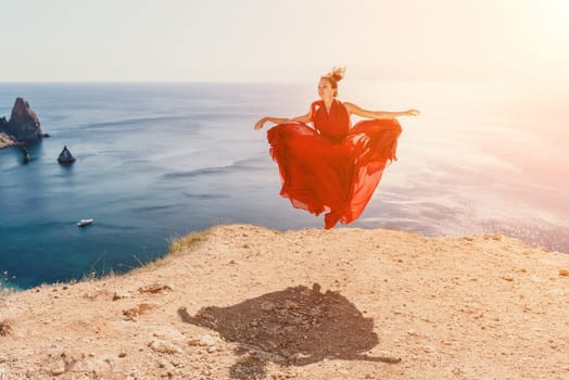 Side view a Young beautiful sensual woman in a red long dress posing on a rock high above the sea during sunrise. Girl on the nature on blue sky background. Fashion photo.