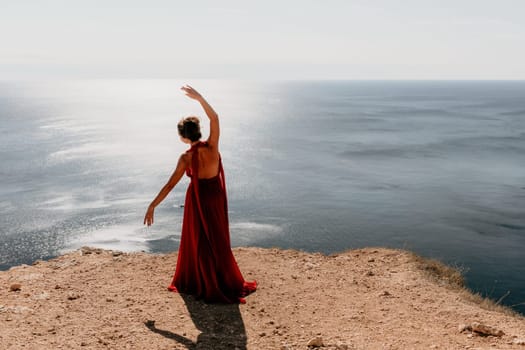 Side view a Young beautiful sensual woman in a red long dress posing on a rock high above the sea during sunrise. Girl on the nature on blue sky background. Fashion photo.