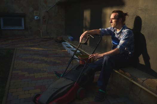 Portrait shot at sunset of tired overworked gardener man, landscaper with electric lawn mower, relaxing after hard working day in the backyard. People. Workman. Labor. Lifestyle. Landscaping industry