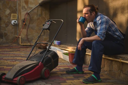 Caucasian professional male worker gardener in blue gardening jumpsuit and checkered shirt taking a break after gardening , drinking coffee from an enamel mug, relaxing outdoor after hard working day