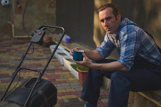 Professional middle aged gardener in blue work jumpsuit and plaid shirt, taking a break after a hard working day in the garden, holding a blue enamel mug of hot coffee, smiling looking at camera