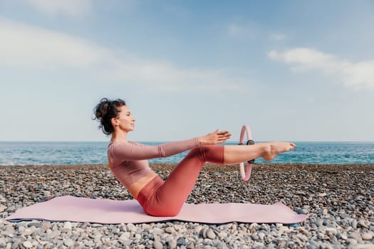 Middle aged well looking woman with black hair doing Pilates with the ring on the yoga mat near the sea on the pebble beach. Female fitness yoga concept. Healthy lifestyle, harmony and meditation.