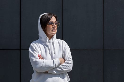 Portrait Mature Brunette Woman With Hood On Head, Wears White Hoodie and Sunglasses With Crossed Arms, Dark Background Wall. Sportive Confident 40 Yo Beautiful Caucasian Woman Outdoor. Horizontal.