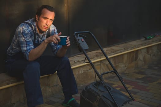 Attractive gardener man in blue work jumpsuit, looking at camera, sitting near a lawn mower with cup of coffee in hands, relaxing after hard working day in the garden. Sunset sun rays fall on his face