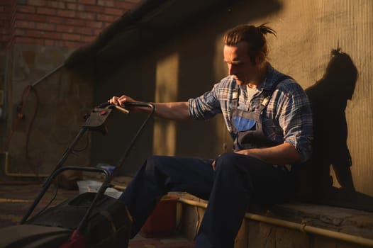 Portrait of overworked gardener man, homeowner, landscaper in blue gardening overalls and checkered shirt, with a lawn mower in the garden at sunset, relaxing after a hard day's work. People. Labour