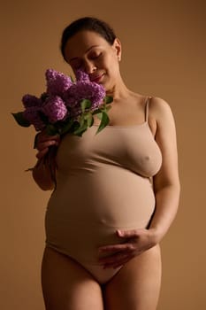 Studio portrait gorgeous adult pregnant woman, expecting baby, caressing her belly, holding bunch of lilacs, isolated beige background. 40 weeks of happy carefree pregnancy. Body positivity. Maternity