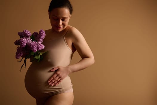 Attractive pregnant woman smiling, touching her belly, enjoying happy moments of her pregnancy, posing with a bunch of purple blooming lilacs, dressed in beige lingerie, isolated on studio background