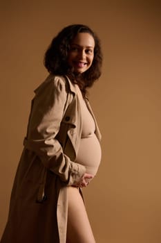 Fashion beauty portrait of expectant gravid mother, elegant pregnant woman in beige lingerie under a trendy trench coat, holding hands on her belly, smiling looking aside. Beautiful pregnancy concept