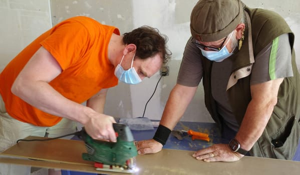 A man saws a board with a jigsaw. A second man holds the board. They wear masks against the dust. This is part of laying a laminate floor