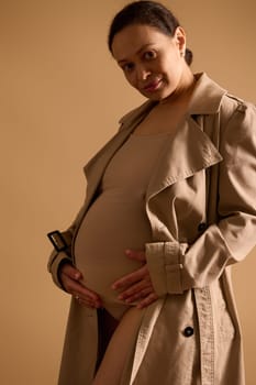 Confident adult gorgeous pregnant woman in elegant trench coat under beige lingerie, strokes her belly, smiles looking at camera, isolated over studio cream background. Beautiful happy pregnancy