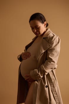 Vertical studio portrait of a beautiful ethnic pregnant woman in beige lingerie and trench coat, gently caressing her big belly, feeling baby kicks, isolated background. Maternity. Pregnancy fashion.