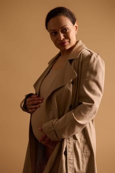 Confident multi-ethnic gravid woman dreamily looking away, touching her pregnant belly, feeling baby kicks, isolated beige background. Happy carefree pregnancy. Mothers and Women's Day concept