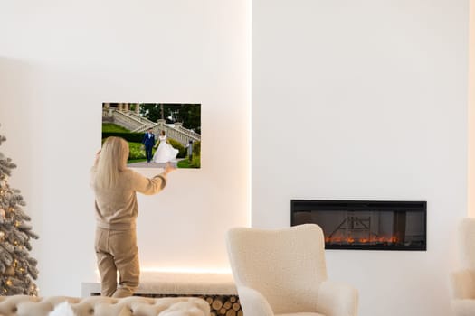 Modern Home Interior And Domestic Decor. Smiling young woman hanging painting, putting photo picture frame on the wall. Casual lady taking care of coziness in her new stylish apartment, profile.