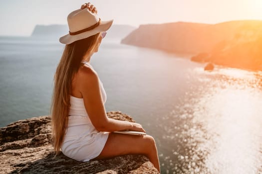 Successful business woman in yellow hat working on laptop by the sea. Pretty lady typing on computer at summer day outdoors. Freelance, travel and holidays concept.