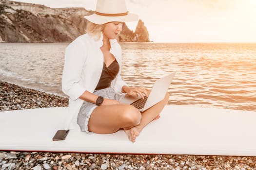 Successful business woman in yellow hat working on laptop by the sea. Pretty lady typing on computer at summer day outdoors. Freelance, travel and holidays concept.