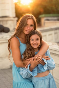 Portrait of mother and daughter in blue dresses with flowing long hair against the backdrop of sunset. The woman hugs and presses the girl to her. They are looking at the camera