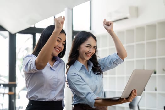 Two young businesswomen show joyful expression of success at work smiling happily with a laptop computer in a modern office.