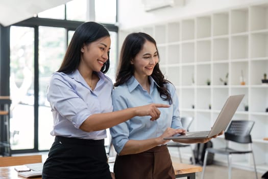 Two Asian business women working together using laptop discussing new startup project ideas. Analyze the planning and statistics of the financial and investment markets at the office.