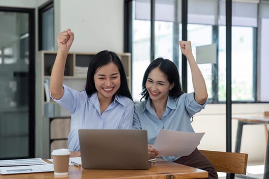 Two young businesswomen show joyful expression of success at work smiling happily with a laptop computer in a modern office.