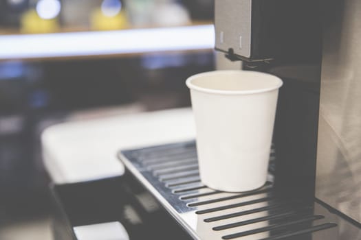 A white paper cup stands on the coffee machine stand, cozy atmosphere of a coffee shop, selective soft focus.