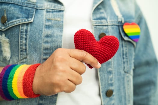 Asian lady wearing rainbow flag wristbands and hold red heart, symbol of LGBT pride month celebrate annual in June social of gay, lesbian, bisexual, transgender, human rights.