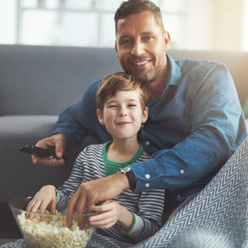 Some popcorn for the movies. Portrait of a carefree young boy and his father watching a movie together while being seated on the floor and eating popcorn at home during the day