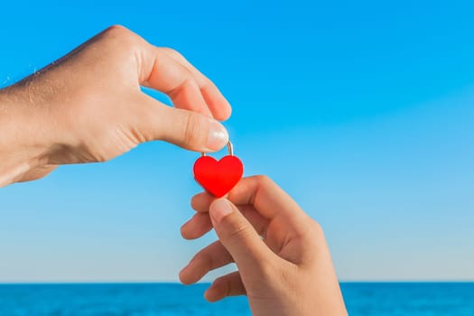 A sign and symbol of love and relationship concept. The hands of a man and a woman hold together a red heart against the blue sea, sky and horizon line.