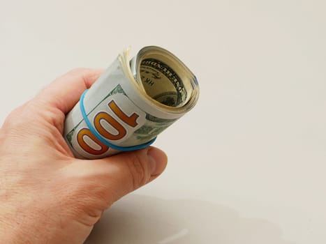 Hand holding a roll of hundred dollar bills with a blue rubber band on a white background.