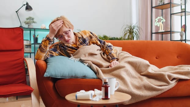 Young ill woman suffering from cold flu or allergy sitting on sofa in living room at home drinking hot tea. Sick blonde girl wrapped in plaid blows sneezes wipes snot into napkin. Quarantine pneumonia