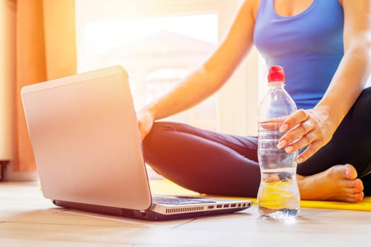 A sporty woman in sportswear is sitting on the floor with a bottle of water and is using a laptop. Sport and recreation concept.