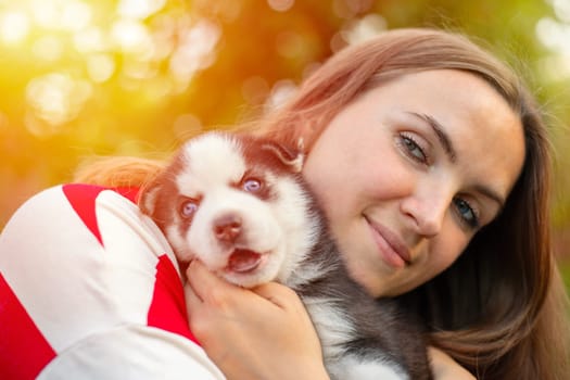 Beautiful woman in a striped t-shirt tenderly hugs a small husky puppy in the park outdoors.