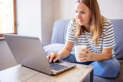 Young attractive woman using laptop computer and drinking morning cup of coffee while wearing a jeans and t-shirt. Brunette female sitting in a living room. Freelancer working from home.