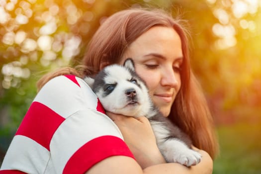 Beautiful woman in a striped t-shirt tenderly hugs a small husky puppy in the park outdoors.