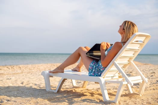 Beautiful young woman working on laptop when sitting in chaise-lounge on sandy beach.