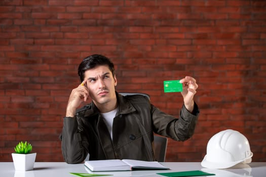 front view male engineer sitting behind his working place and holding green credit card builder money plan corporate contractor business property