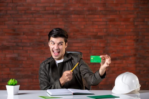 front view male engineer sitting behind his working place and holding green credit card builder plan corporate contractor business property agenda