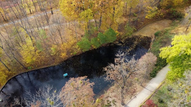 Flying over autumn park. Many trees with yellow green and fallen leaves, lakes, people walking dirt paths in park on sunny autumn day. Top view. Aerial drone view. Natural background