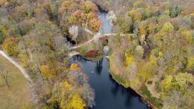 Beautiful scenery view of an autumn park with trees with yellow fallen leaves, lakes, architecture, glades and people walking along dirt paths on an autumn day. Flying over the autumn park. Top view.