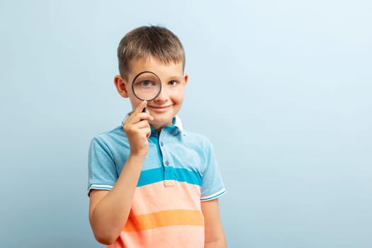 Curious schoolboy in casual clothes looking through the magnifying glass on blue background.