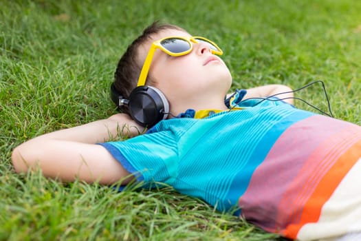 Happy boy in sunglasses lying on the grass and listening to music in headphones on a sunny day.