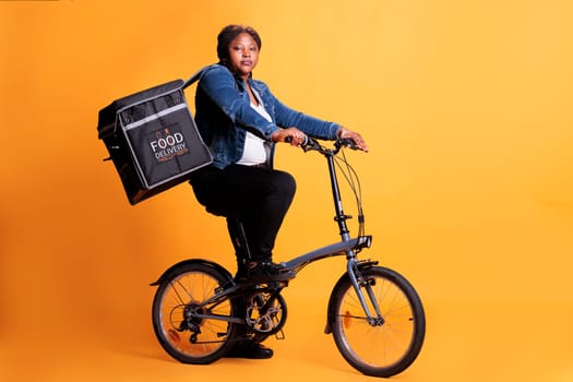 Food delivery employee riding bike while carrying takeaway thermal backpack ready to deliver order to client during lunch time, standing over yellow background. Food service and transportation