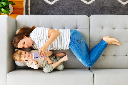 Woman and her little son lying on cozy sofa with smartphone, above view. Family spend leisure together at home using smartphone, play games, watch video, take selfies