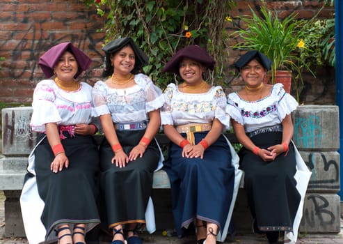 Front view of four Latin women sitting on a bench in traditional clothing from their country looking at the camera and smiling.
