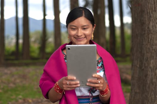 Front view of a happy a young Latin woman in traditional clothing from her country in the forest using a digital tablet.