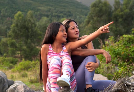 Front view of a happy daughter pointing something to her mother in nature sitting in the mountains.