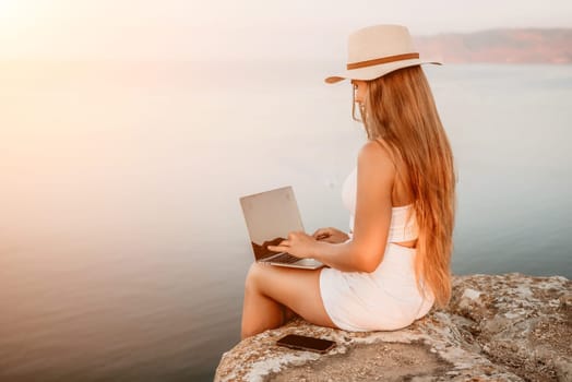 Digital nomad, Business woman working on laptop by the sea. Pretty lady typing on computer by the sea at sunset, makes a business transaction online from a distance. Freelance, remote work on vacation