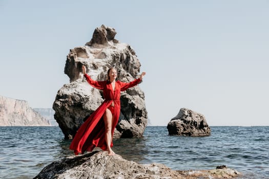 Woman travel sea. Young Happy woman in a long red dress posing on a beach near the sea on background of volcanic rocks, like in Iceland, sharing travel adventure journey