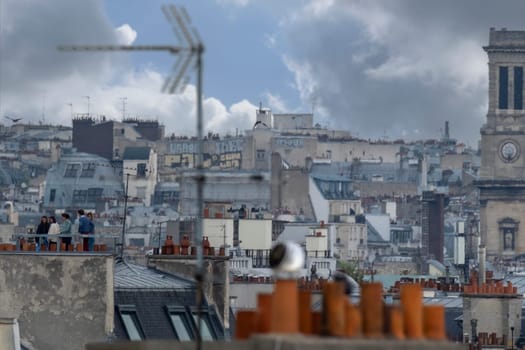 Selective focus aerial view of rooftops in Paris showing clay chimney pots, gothic tower, TV antennae and people on a rooftop terrace. Telephoto shot.