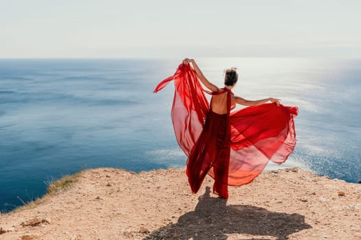 Side view a Young beautiful sensual woman in a red long dress posing on a rock high above the sea during sunrise. Girl on the nature on blue sky background. Fashion photo.