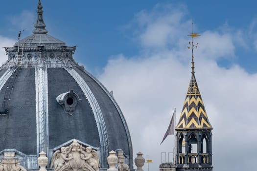 A dome with carved stone ornamentation and a tower with yellow and black roof tiles and a spire. Telephoto lens close up of architectural details in Paris.